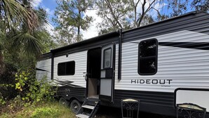 A HIDEOUT RV with a sliding door, a small porch area with two chairs, and a step-up ladder leading to the door.