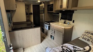 A compact RV interior with a kitchenette, a bed with a 'Welcome' mat, and a 'Let the adventure begin' pillow.