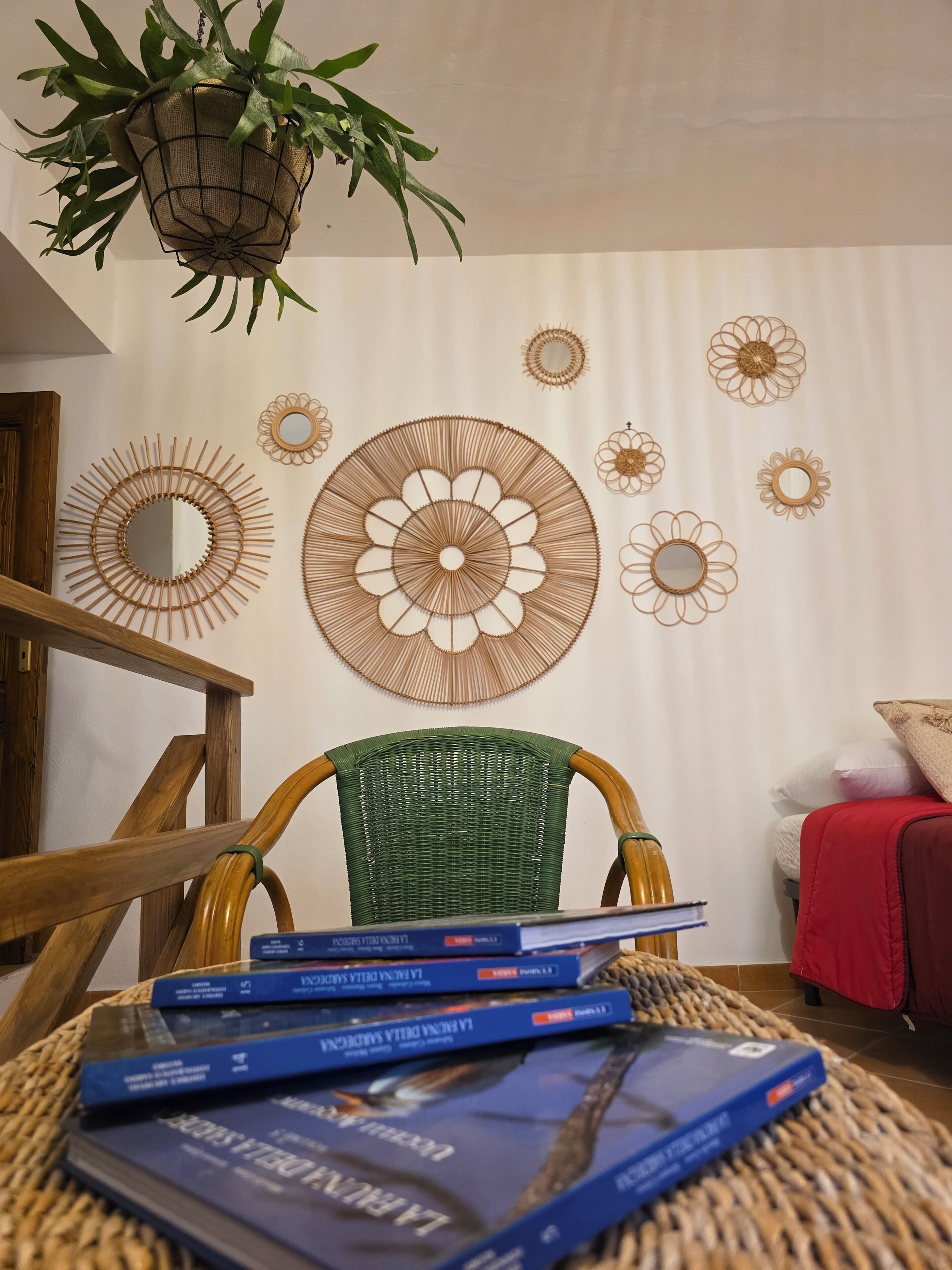 Una stanza con una poltrona verde intrecciata, una sedia di legno e un divano rosso. Sono presenti specchi decorativi a parete e una pianta pensile.