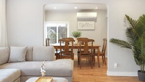 Een moderne woonkamer met een grijze bank, een houten eettafel en stoelen. Er staat een potplant en een lijst met een citaat aan de muur.
