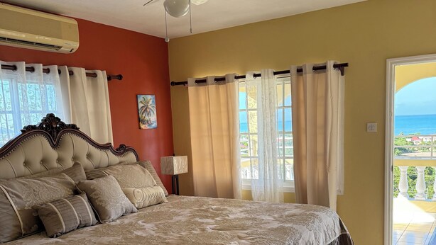 A bedroom with a large bed, a ceiling fan, and a view of the ocean through the window. - Superstar Living : Beautiful 3-Bed Room Suite with Balcony and Sea View (Treasure Beach)