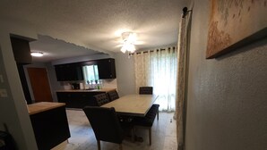 A dining area with a marble floor, a wooden table, and black upholstered chairs. There is a kitchen in the background with dark cabinets and a window with patterned curtains. - Relaxing Ranch (sodus)