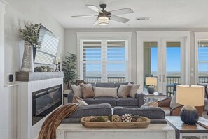 Ein Wohnzimmer mit Kamin, einer grauen Sofagarnitur, Deckenventilator und großen Fenstern mit Jalousien.