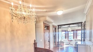 A grand staircase with a wooden handrail leading to a landing. A chandelier hangs from the ceiling, and a large window provides natural light.