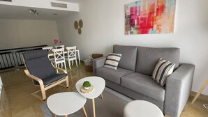 Una sala de estar moderna con un sofá gris, un sillón oscuro y una pequeña mesa blanca. Al fondo, un comedor con sillas blancas y una mesa.