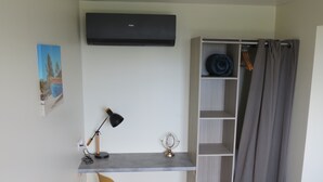 Ein kleines Zimmer mit Schreibtisch, Stuhl, Stehlampe, einem kleinen Tisch mit Dekoration und einem Kleiderschrank mit Regalen.