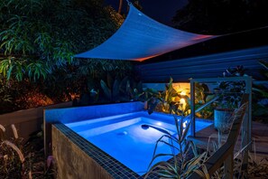 Un jacuzzi con interior iluminado en azul, rodeado de plantas y una terraza de madera.