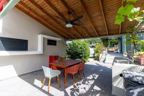 Un patio exterior con una mesa y sillas de madera, un ventilador de techo y vistas del jardín circundante.