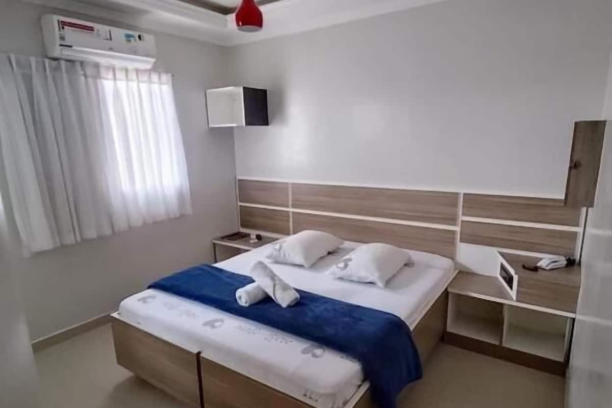 Habitación de hotel moderna con una cama, mesitas de noche, una lámpara colgante roja, un equipo de aire acondicionado y una ventana con cortinas.