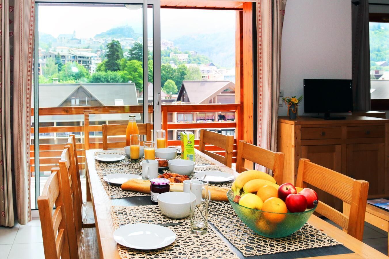 Una mesa de comedor con alimentos para el desayuno, un cuenco de fruta y vistas a un paisaje urbano a través de amplios ventanales.