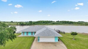 A two-story house with a garage, a grassy yard, and a view of a lake. - Matagorda-Stunning Waterfront Views-Swim Spa-Private Pier-Relax (Matagorda)