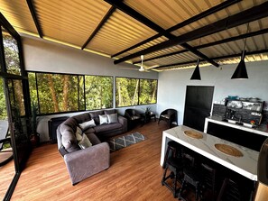 A modern living room with a sectional sofa, a dining area with bar stools, large windows with a forest view, and a kitchen with black and white tiles. - Casa de Luna Villa with Ocean View (Uvita)