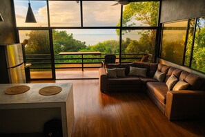 Ein modernes Wohnzimmer mit großem Fenster, einem Ecksofa, einem Holztisch und einer Pendelleuchte.