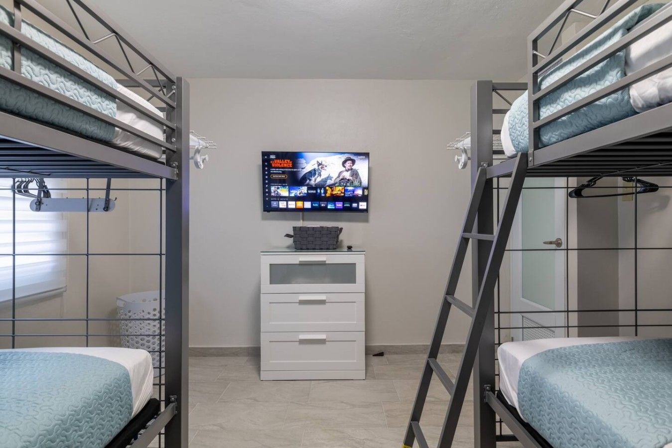 A bunk bed room with a television mounted on the wall, a small white dresser, and a ladder.