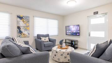 Een woonkamer met grijze banken, een grijze fauteuil, een salontafel met een decoratief object, een flatscreen televisie aan de muur en een deur met een 'EXIT'-bord.