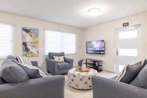 Una sala de estar con sofás grises, un sillón gris, una mesa de centro con un objeto decorativo, un televisor de pantalla plana montado en la pared y una puerta con un letrero que dice 'EXIT'.
