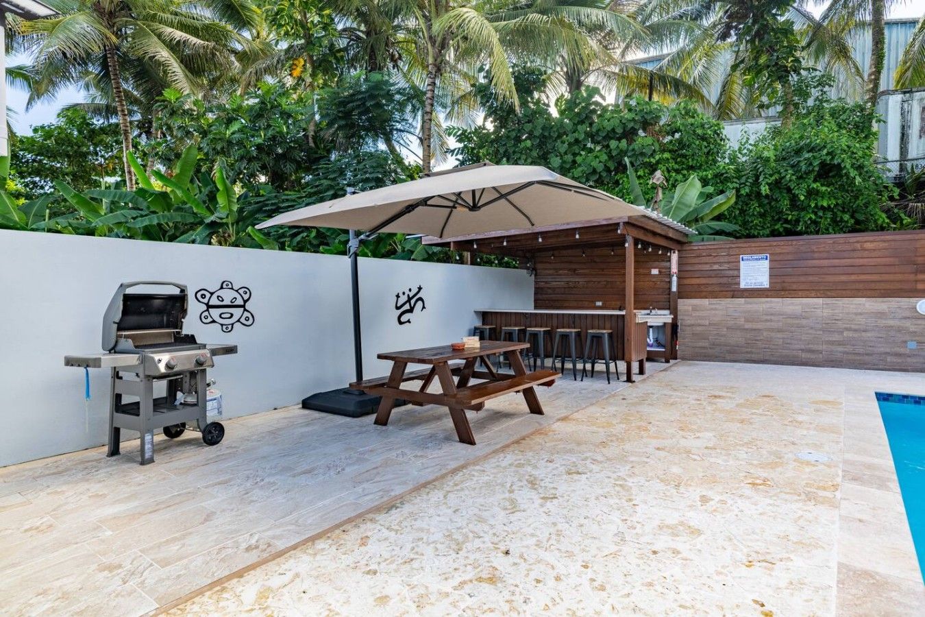 An outdoor patio with a barbecue grill, picnic table, and umbrella, surrounded by a pool and lush greenery.