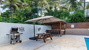 Une terrasse extérieure équipée d’un barbecue, d’une table de pique-nique et d’un parasol, le tout entouré d’une piscine et d’une végétation luxuriante.