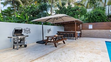 An outdoor patio with a barbecue grill, picnic table, and umbrella, surrounded by a pool and lush greenery.