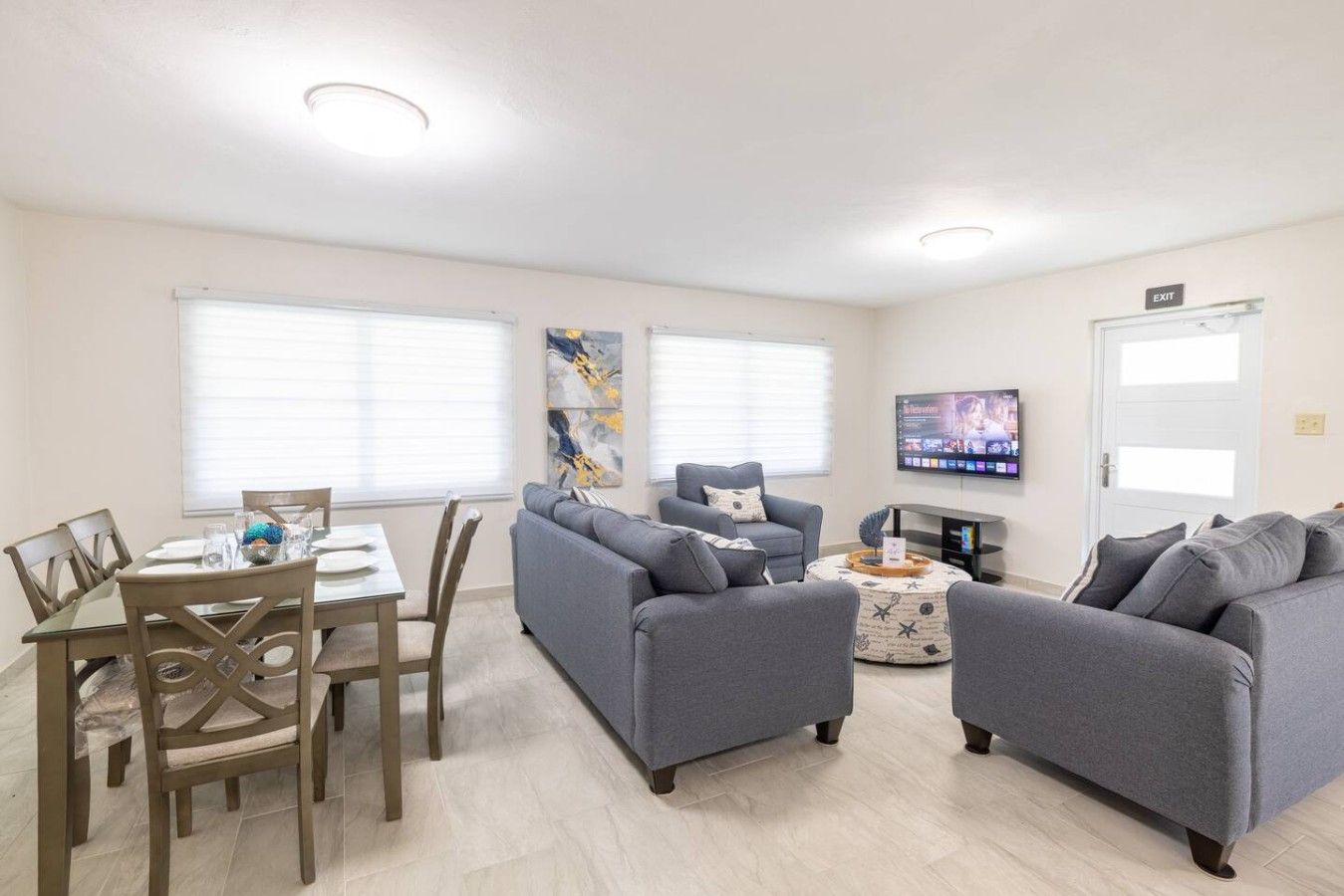 A modern living room with a dining area, grey sofas, a coffee table, and a flat-screen TV mounted on the wall.