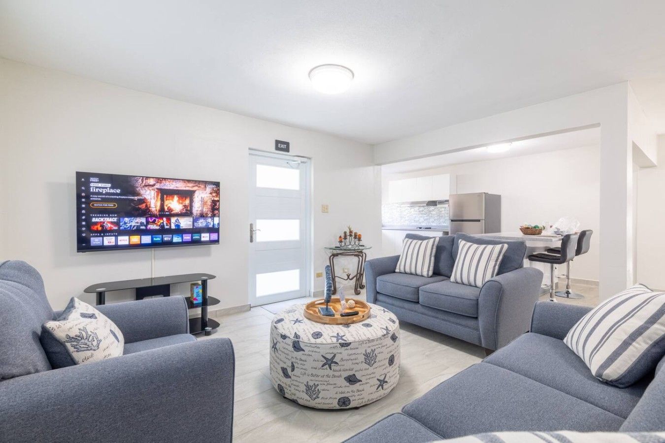 Ein modernes Wohnzimmer mit Flachbildfernseher, grauen Sofas, einem Couchtisch und einer Küchenzeile im Hintergrund.