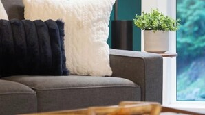 Een moderne woonkamer met een grijze bank, een zwart-wit kussen met textuur, een vloerlamp, een kleine potplant en een houten dienblad op een donkere tafel.