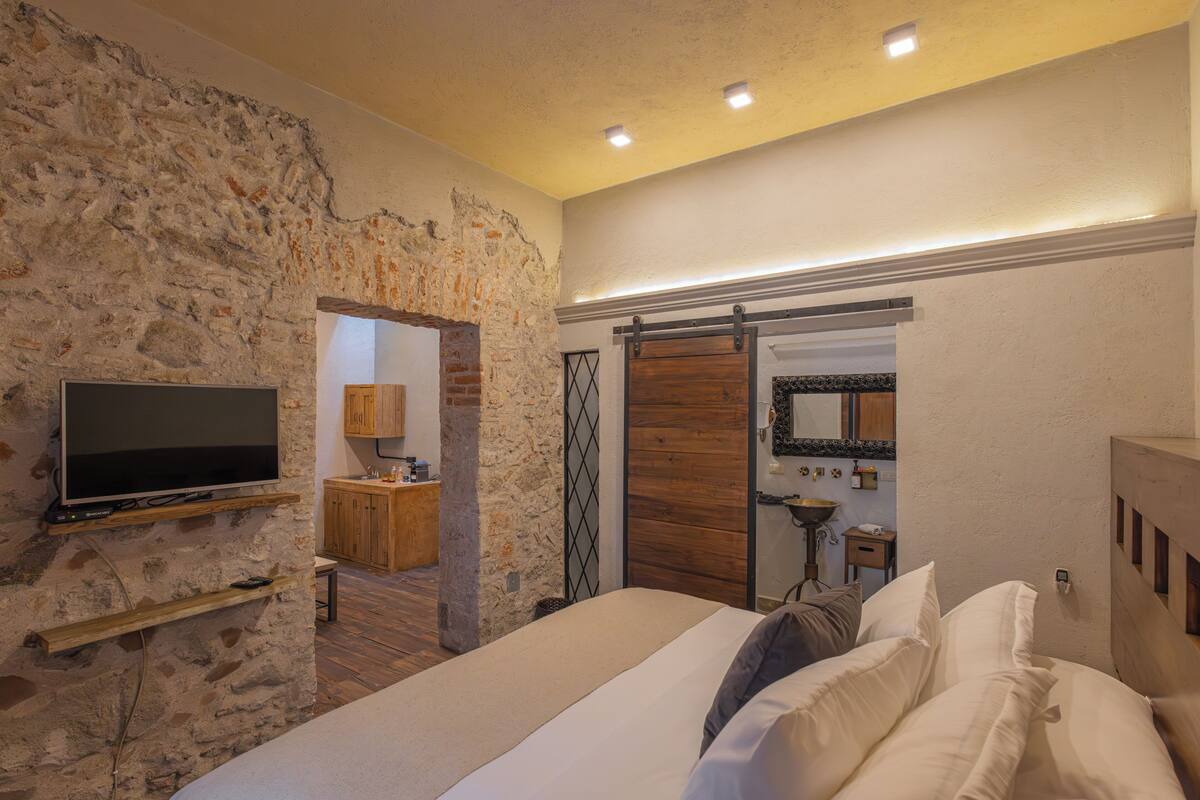 A bedroom with a stone wall, a bed with white linens, a wooden wardrobe, and a mounted television.