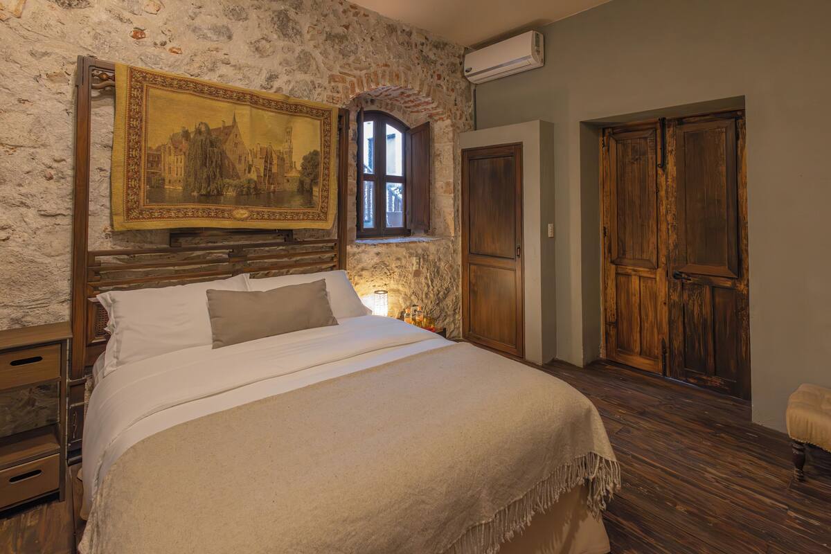 A bedroom with a stone wall, a bed with white linens, a wooden headboard, a large framed painting, and a window with wooden shutters.