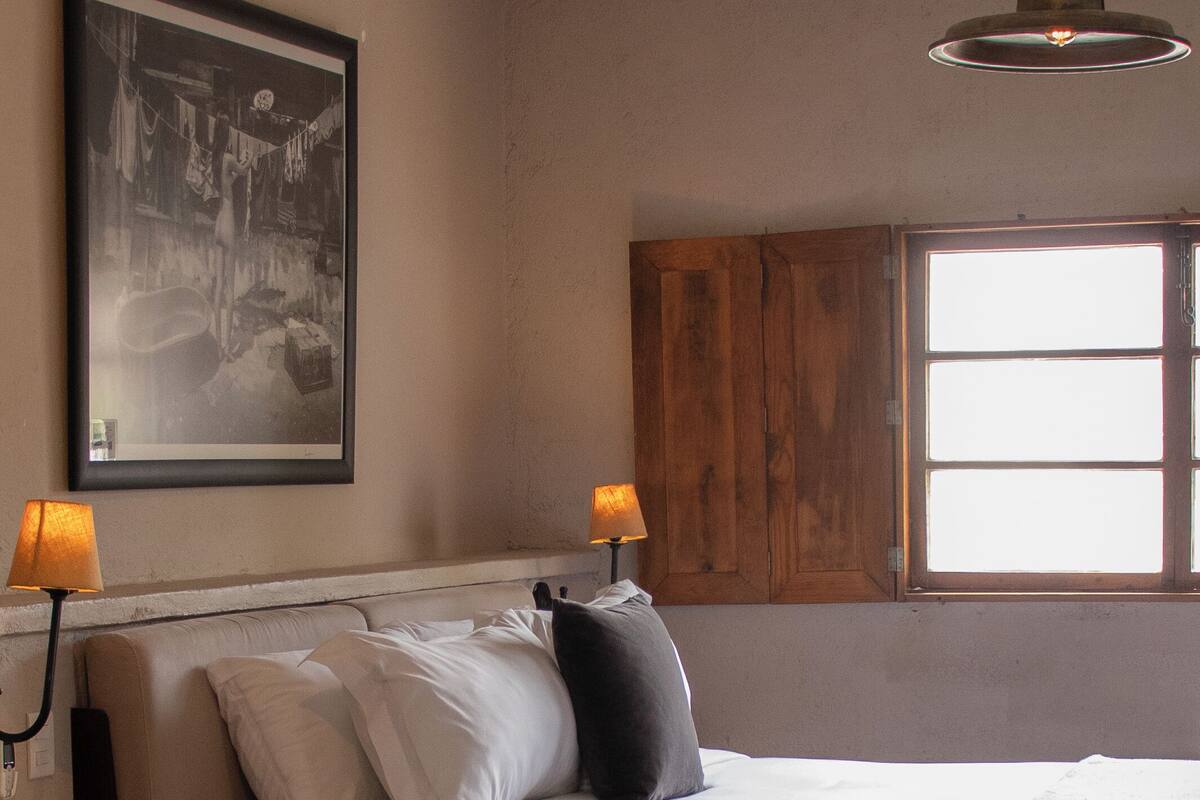 A bedroom with a bed, two bedside lamps, a wooden cabinet, a framed picture, and a window.