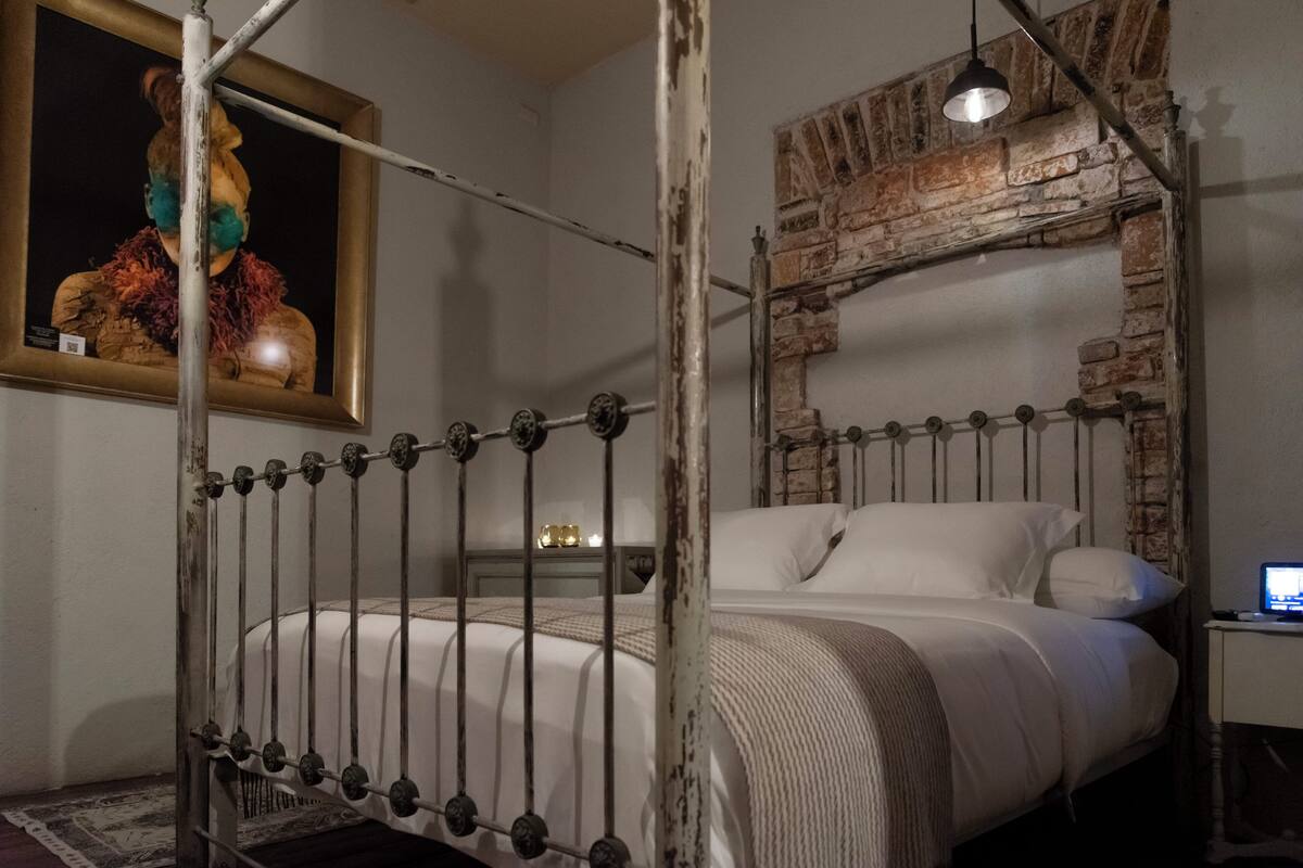 A bedroom with a metal bed frame, a brick wall, a wooden headboard, a bedside table with a lamp, and a painting on the wall.