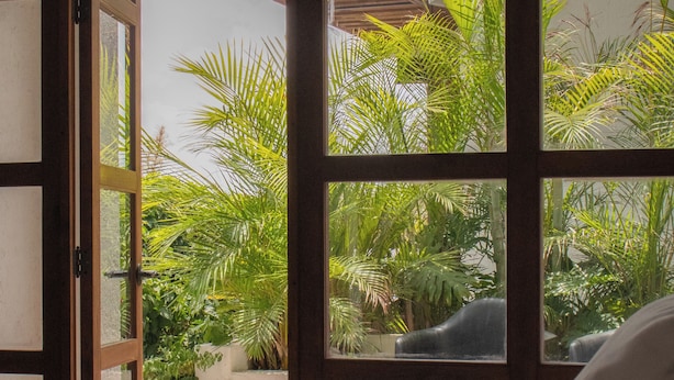 A bedroom with a bed, a view of lush greenery through large windows, and a chair outside. - Hotel C. Andreu  (San Miguel de Allende)