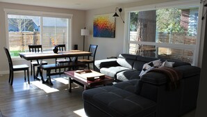 Una sala de estar con un sofá de color oscuro, una mesa de comedor de madera con sillas y un ventanal con vista a un patio trasero.