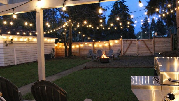 Un área de patio al aire libre con luces colgantes, un hogar para fogata y una parrilla.