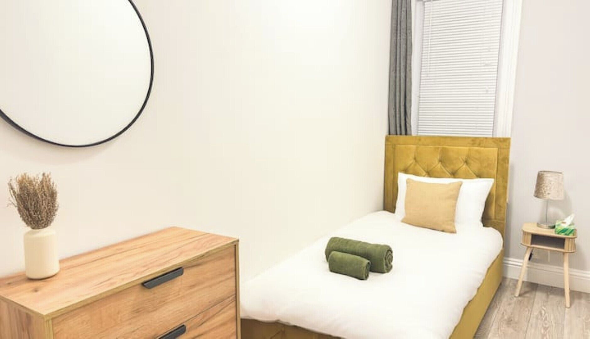 Un dormitorio ordenado con cama, mesita de noche, lámpara y cómoda.