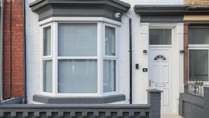 A corner bay window with shutters, a white door with a number plate, and a stone wall.