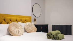 A neatly made bed with two large pillows, a black nightstand with a mirror, and a rolled green towel.