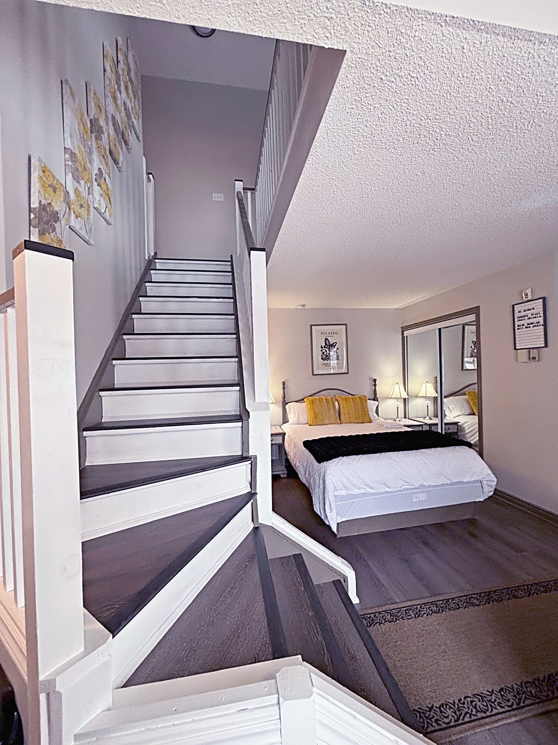 A bedroom with a bed, bedside tables, a nightstand, a mirror, and a staircase leading to another room.