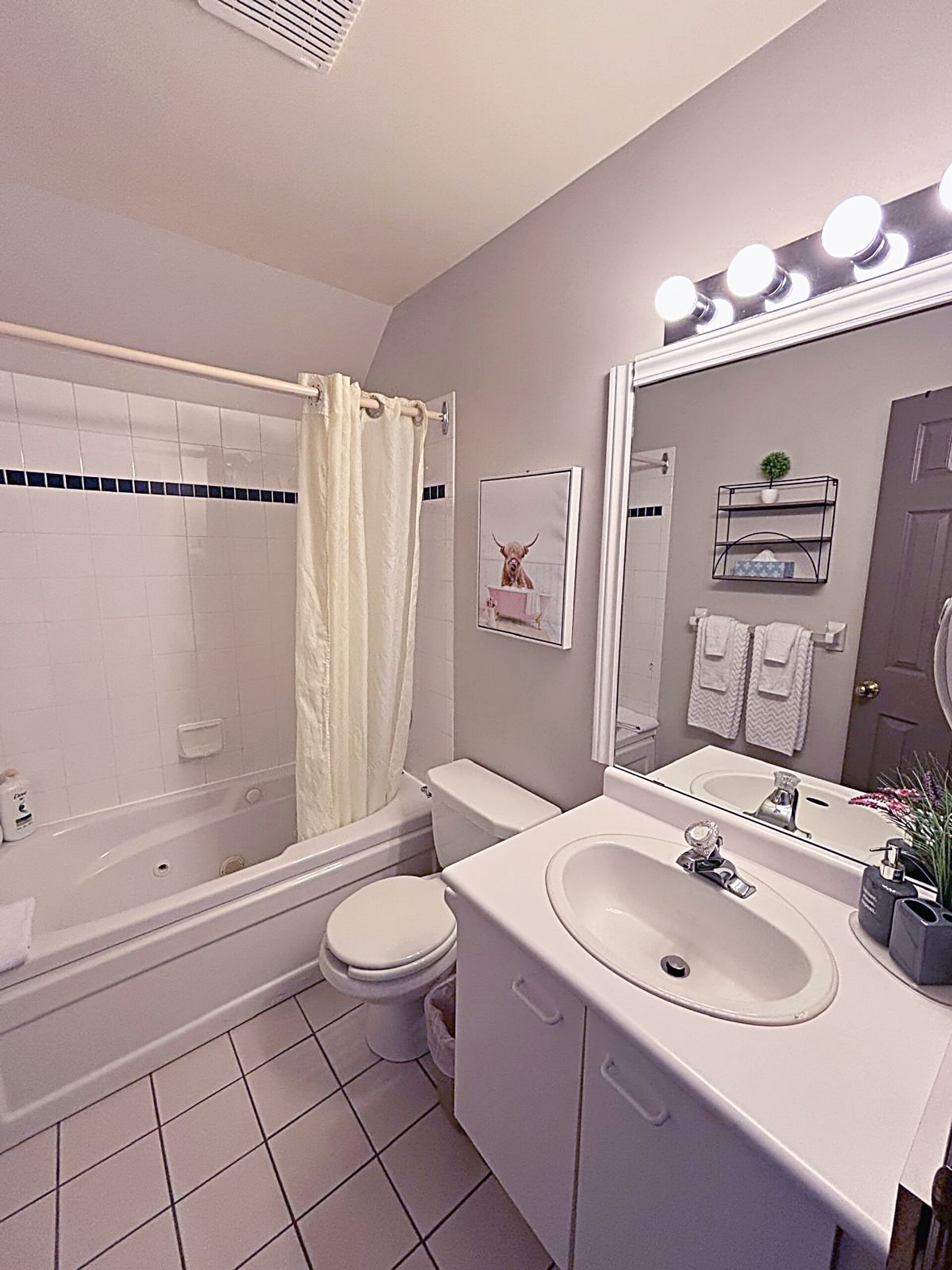 Ein Badezimmer mit Badewanne, Toilette und Waschbecken. Ein Duschvorhang, ein Spiegel mit Beleuchtung und eine kleine Pflanze auf dem Waschbecken sind vorhanden.