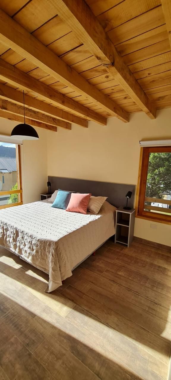 A bedroom with a wooden ceiling, a bed with a white bedspread, two pillows, a nightstand, and a window with a view of trees.