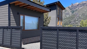 Una casa moderna con un amplio ventanal, una pérgola de madera y vistas a las montañas.