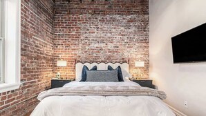 A bedroom with a brick wall, a bed with white bedding, two bedside tables with lamps, and a mounted TV.