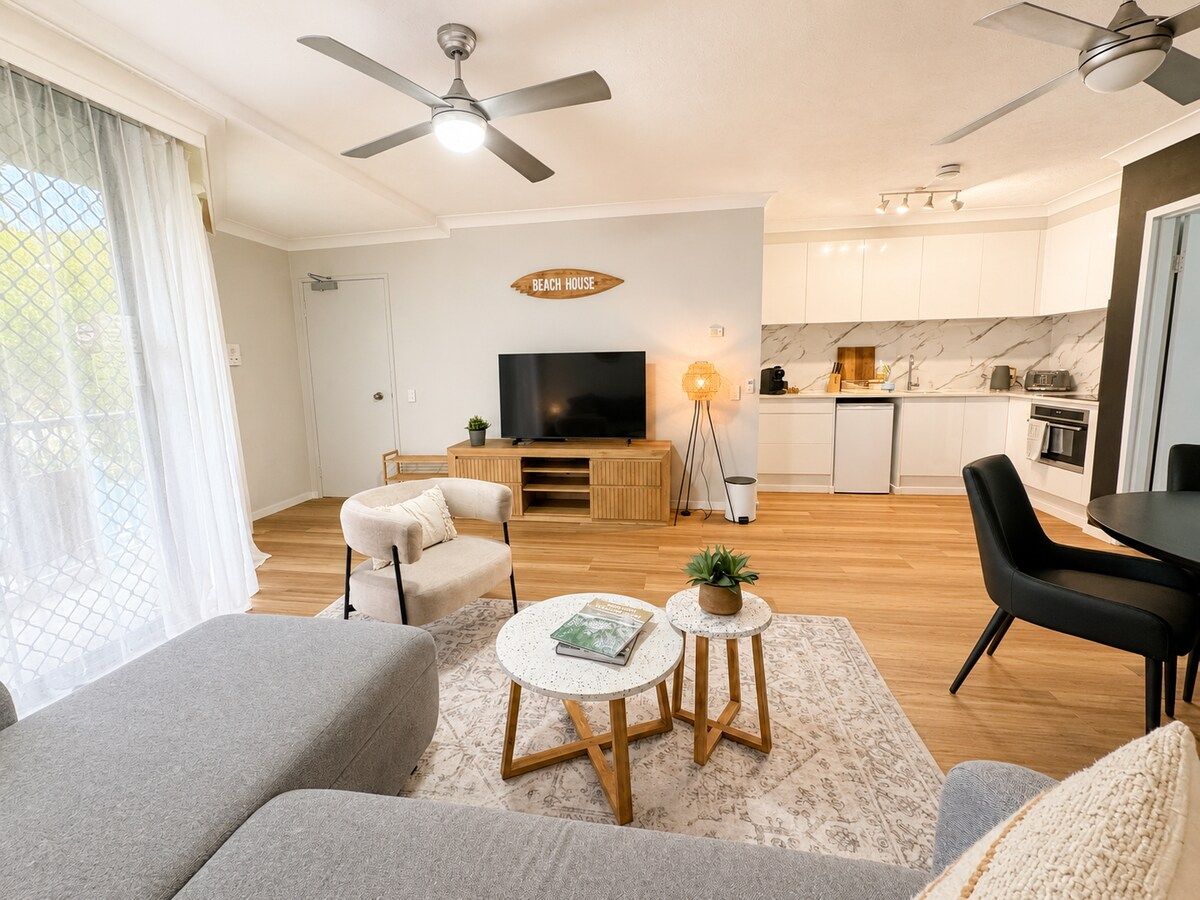 A modern living room with a sofa, coffee table, and TV. There is a kitchen in the background with white cabinets and a dining area.