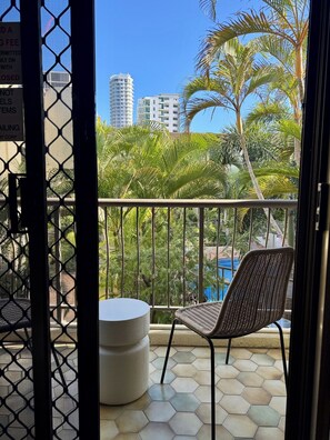 Un balcon avec un fauteuil en osier et une table ronde, donnant sur une piscine et des palmiers, avec des bâtiments en arrière-plan.