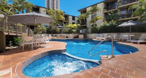 Une piscine entourée de chaises longues et de parasols, elle-même bordée d’un immeuble à plusieurs étages doté de balcons.