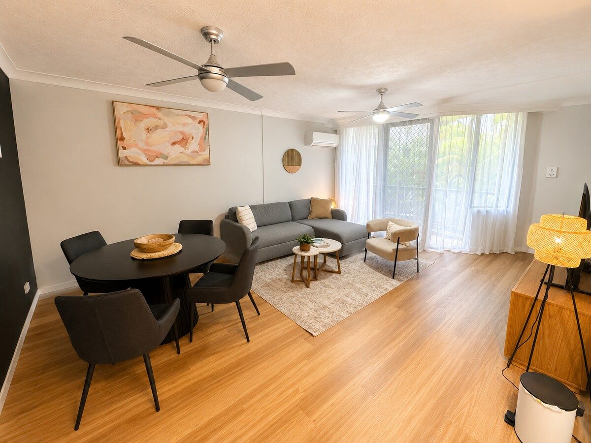 A modern living room with a grey sofa, a dining table with chairs, a ceiling fan, and a large window with sheer curtains.