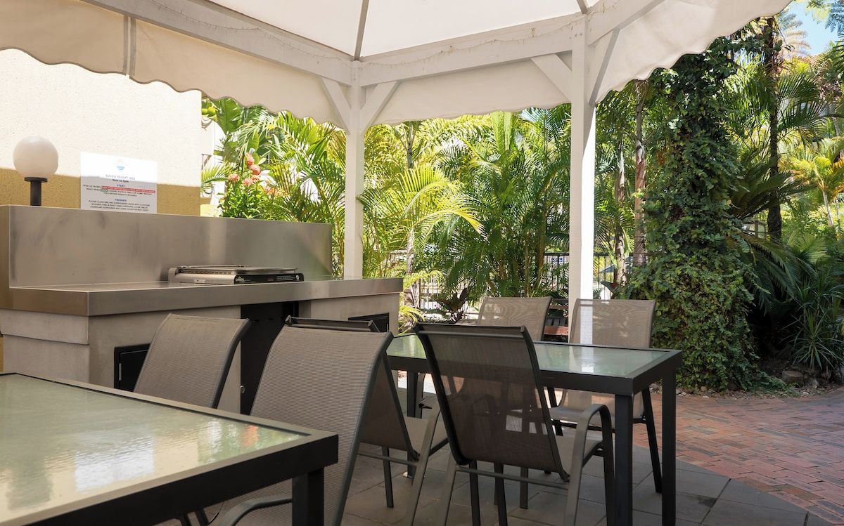 An outdoor patio area with tables and chairs, a grill, and lush greenery in the background.
