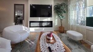 Una sala de estar moderna con chimenea, un televisor de pantalla plana grande, un otoman blanco, una mesa de centro de madera con un control remoto y una planta en maceta.