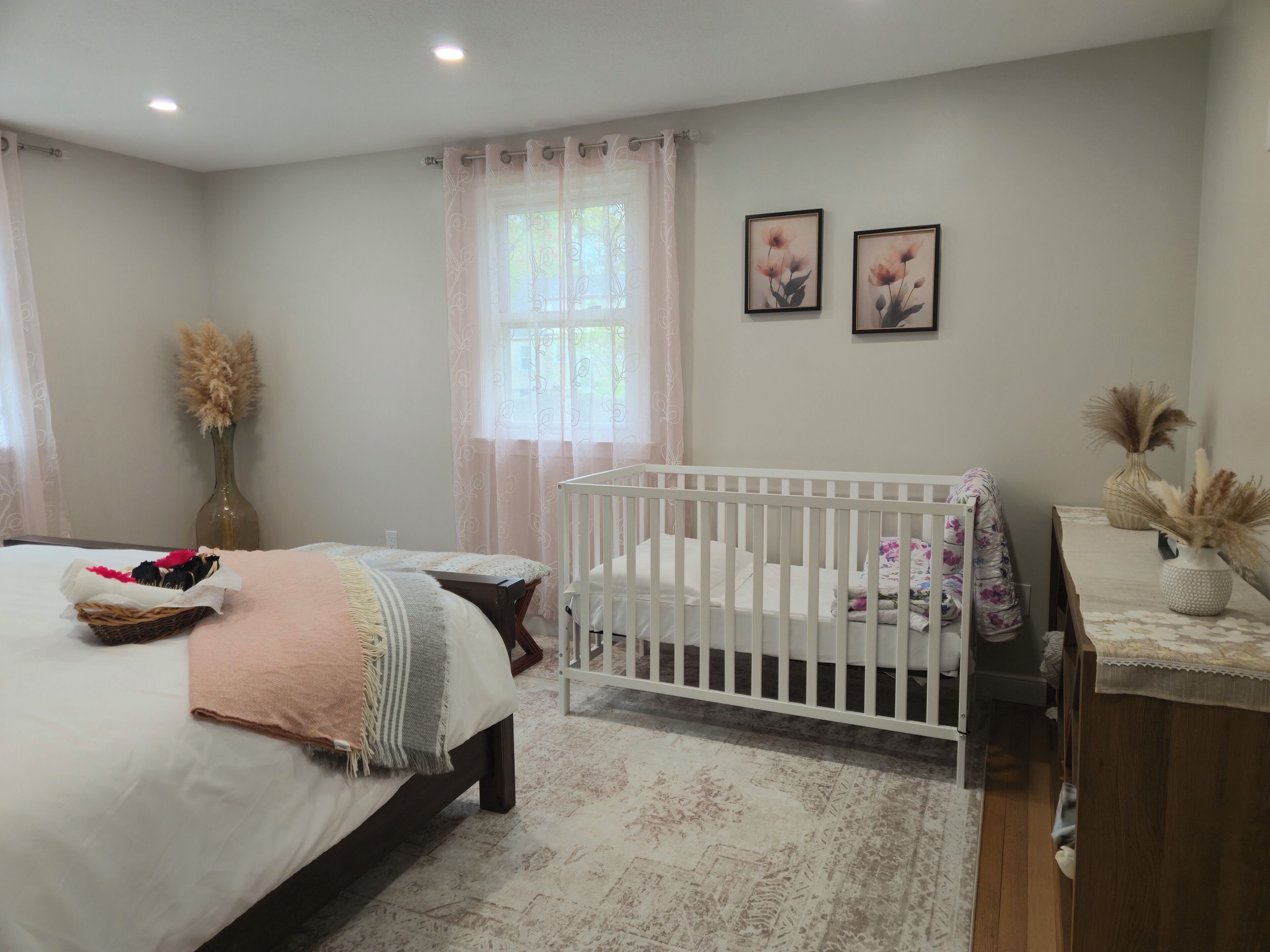Ein Schlafzimmer mit einem Babybett, einem Bett mit einem Korb voller Gegenstände, einer Vase mit getrockneten Blumen und zwei gerahmten Bildern an der Wand.