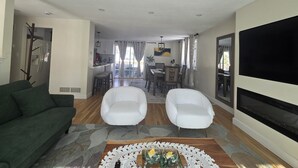 Sala de estar con sofá verde, dos sillones blancos, una mesa de centro con adornos, televisor de pantalla plana y zona de cocina con refrigerador y mesa de comedor.