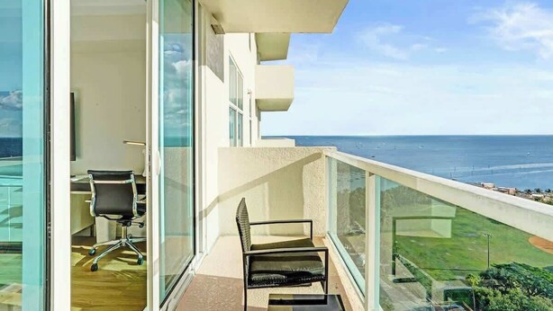 A balcony with a view of the ocean, a chair, and a desk with a chair. - Coconut Grove Suite | 20th Floor Bay Views (Miami)